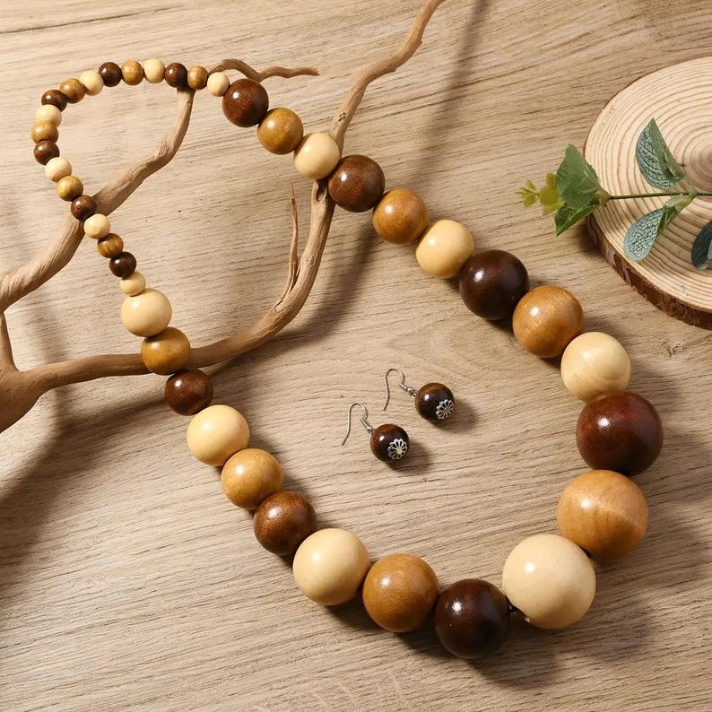 May include: A wooden bead necklace and matching earrings set. The necklace features large, round beads in shades of light tan, medium brown, and dark brown. The earrings have a small, round bead with a silver-colored accent.