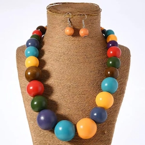 Puede incluir: Un conjunto de collar y pendientes coloridos. El collar presenta grandes cuentas redondas en varios colores, incluyendo rojo, verde, azul, amarillo y marrón. Los pendientes son pequeños, redondos y naranjas. La joyería se exhibe en un busto marrón.