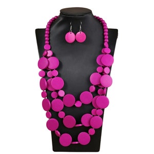 May include: A vibrant fuchsia necklace and earring set displayed on a black mannequin. The necklace features multiple strands of round beads and large, flat, circular wooden pendants. The matching earrings are simple, round discs.