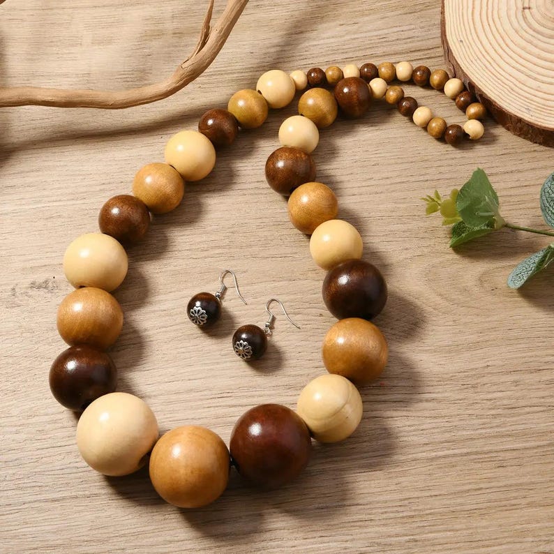 May include: A wooden bead necklace and matching earrings. The necklace features large, round beads in shades of light tan, medium brown, and dark brown. The earrings have dark brown beads with silver-colored decorative accents. The jewelry is displayed on a wooden surface.