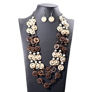 Puede incluir: Un conjunto de collar de tres hilos y pendientes a juego. El collar presenta cuentas de madera de color marrón oscuro, marrón claro y blanco alternadas. Los pendientes son discos de madera redondos y sencillos de color marrón claro. Las joyas se exhiben en un maniquí negro.