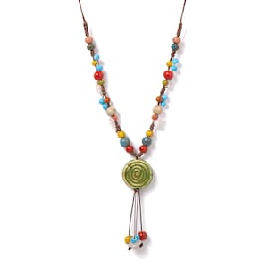 Op de afbeelding: Een handgemaakte ketting met een verscheidenheid aan kleurrijke kralen in rood, blauw, geel en beige. Het middelpunt is een groene keramische hanger met een spiraalontwerp, aangevuld met bungelende kralen. De ketting is geregen op een bruin koord.