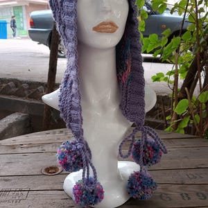 May include: A lavender knitted hat with long ear flaps and three pom-poms. The hat is displayed on a white mannequin head. The pom-poms are a mix of pink, blue, and purple. The hat is made of a soft, textured yarn.