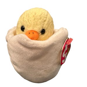 May include: A yellow plush chick emerging from a soft, off-white egg-shaped nest. The chick has black eyes and an orange beak. A red tag is attached to the nest.