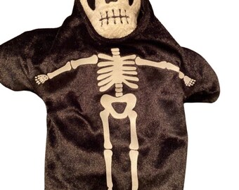 May include: A black plush skeleton puppet with a white skeleton design on the front. The puppet has a white skull face with a black hood. The arms are outstretched, and the legs are visible at the bottom.