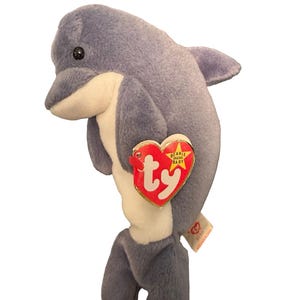 Puede incluir: Un peluche de delfín en tonos azules y blancos. El delfín tiene una etiqueta en forma de corazón con la palabra "ty" en rojo. El juguete es un artículo de colección.