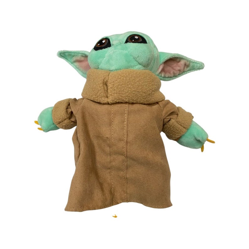 May include: A plush toy of a character with large brown eyes, green skin, and large ears. The character is wearing a tan coat with a brown collar. The toy has small green hands and is set against a white background.