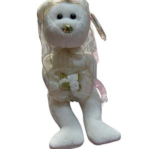 May include: White plush bear wearing a white veil with gold trim and holding a bouquet of white flowers. The bear has a gold nose and black eyes.