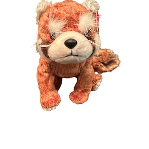 May include: A plush red panda toy with orange and brown fur, white accents around the eyes and ears, and a black nose. The stuffed animal is in a seated position, with its paws visible. A red tag is attached to the ear.