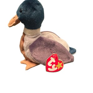 May include: A plush mallard duck toy with a teal head, tan beak, and purple and brown wings. The toy has a red heart-shaped tag attached. The tag reads "Beanie Baby".