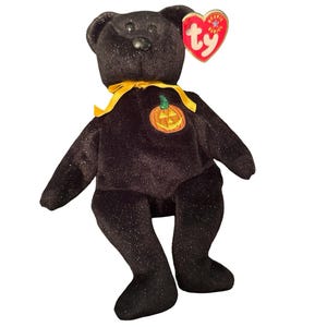 May include: A black plush bear with a subtle glittery finish, featuring a yellow ribbon tied around its neck and an embroidered orange pumpkin on its chest. A heart-shaped red and gold Ty Beanie Babies tag is attached to the bear's left ear.