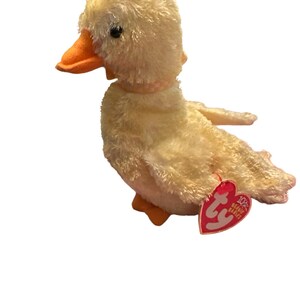 May include: A yellow plush duck with an orange beak and feet. The duck has a pink collar and a Ty Beanie Babies tag attached to its wing. The tag is red with white text that says "10th Beanie Babies".