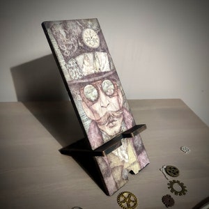 May include: A wooden phone stand with a steampunk design. The stand features a vintage illustration of a man wearing goggles and a top hat. The stand is decorated with gears and other steampunk elements.