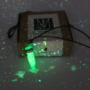 Puede incluir: Un collar con un colgante verde que brilla en la oscuridad, con un diseño retorcido y en espiral. El collar está en un cordón negro y está sentado en una caja marrón con salpicaduras de pintura blanca. La caja tiene el texto "All Handmade" en ella.