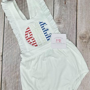 May include: White romper with ruffle trim and embroidered initials in red and blue. The romper has a scoop neckline and a bubble bottom. The tag reads "Creative Thread Embroidery".