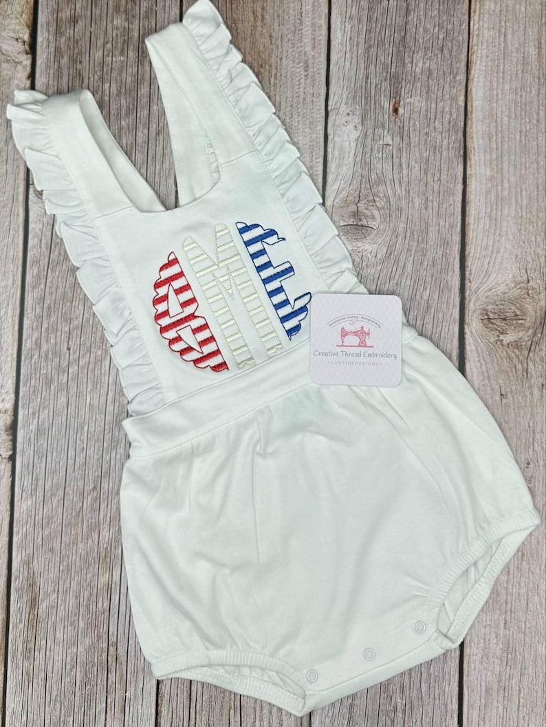 May include: White romper with ruffle trim and embroidered initials in red and blue. The romper has a scoop neckline and a bubble bottom. The tag reads "Creative Thread Embroidery".