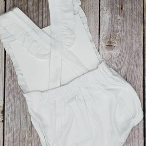 May include: A white baby romper with ruffled straps and detailing. The romper has a gathered waist and a rounded bottom. The fabric appears to be a soft cotton or similar material. The background is a wooden surface.