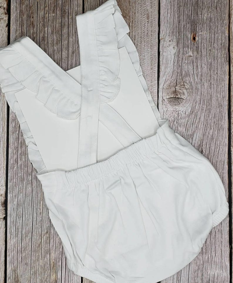 May include: A white baby romper with ruffled straps and detailing. The romper has a gathered waist and a rounded bottom. The fabric appears to be a soft cotton or similar material. The background is a wooden surface.