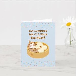 May include: A birthday card with a light blue background and colorful confetti. The card features a cartoon sloth wearing a party hat, nestled in a bamboo steamer with dumplings. Text on the card reads "Dim Sumbody Say It's Your Birthday?"
