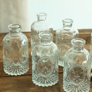 May include: Several clear glass bottles with embossed sun-like patterns. The bottles have a rounded shape and narrow necks. They are displayed on a wooden surface, highlighting their decorative details and vintage style.