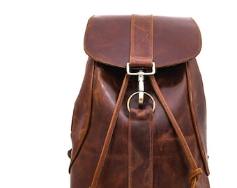 May include: A brown leather backpack featuring a fold-over flap with a central vertical strap. The closure consists of a metallic lobster clasp attached to a circular ring. Two thin leather drawstrings with knotted ends hang from the front.