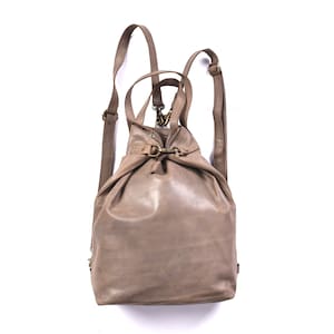 May include: A taupe leather backpack featuring a slouchy, unstructured silhouette with a drawstring closure, antique brass-toned metal hardware, two adjustable shoulder straps, and a top handle for versatile carrying options.