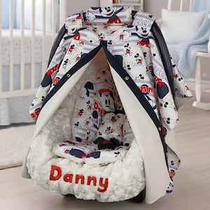 May include: A baby car seat with a custom Mickey Mouse design. The seat features a white, red, and navy blue pattern with the name "Danny" embroidered in red. The seat is covered with a soft, white, plush material.