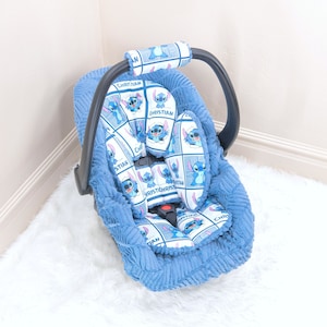 May include: A blue baby car seat cover with a white and blue patterned design featuring a cartoon character. The pattern includes the name "Christian" repeated throughout.