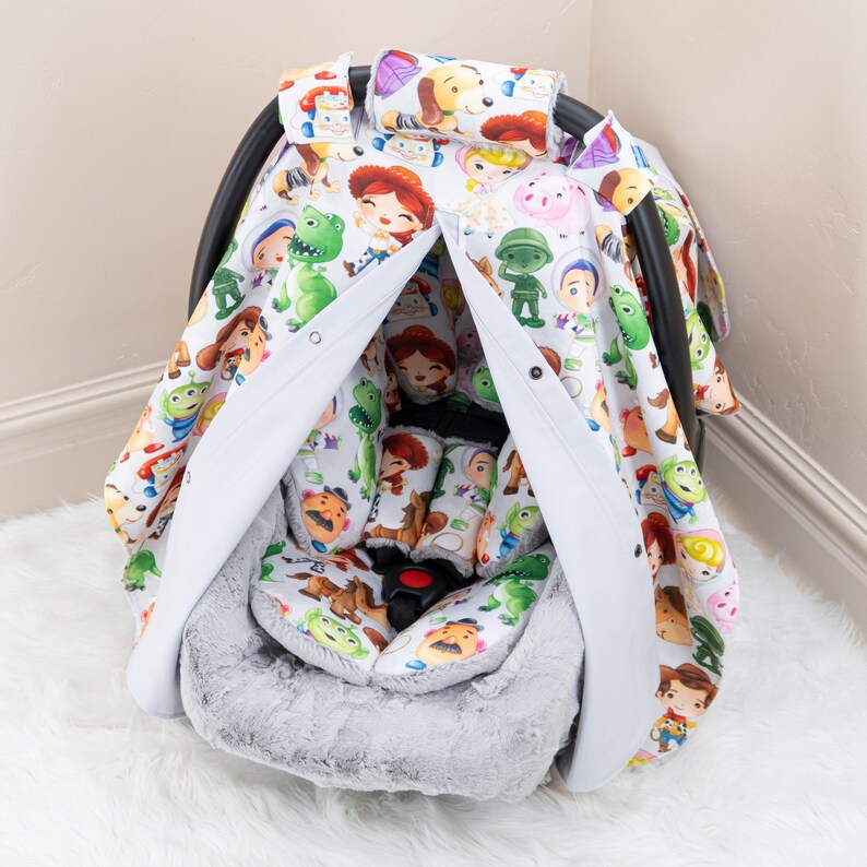 May include: A white car seat cover with a gray faux fur liner. The cover features a Toy Story theme with characters like Woody, Buzz Lightyear, and Jessie printed on it.