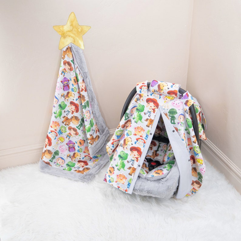May include: A white and grey baby car seat cover with a Toy Story character print. The cover is made of soft, plush fabric and features a variety of characters from the popular Disney movie.