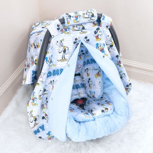 May include: A blue baby car seat cover featuring a Mickey Mouse and Donald Duck print. The cover has a soft, plush lining and a hood that can be pulled up to shield the baby from the sun or wind.