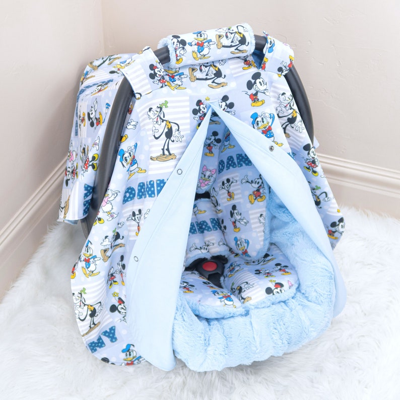 May include: A blue baby car seat cover featuring a Mickey Mouse and Donald Duck print. The cover has a soft, plush lining and a hood that can be pulled up to shield the baby from the sun or wind.