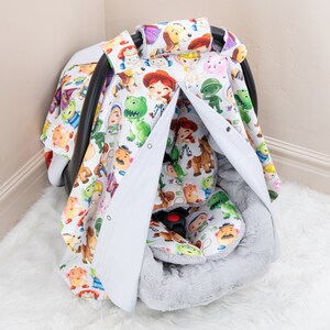 May include: A white car seat cover with a Toy Story print featuring characters like Woody, Buzz Lightyear, and Jessie. The cover has a gray faux fur lining and a white zippered flap.