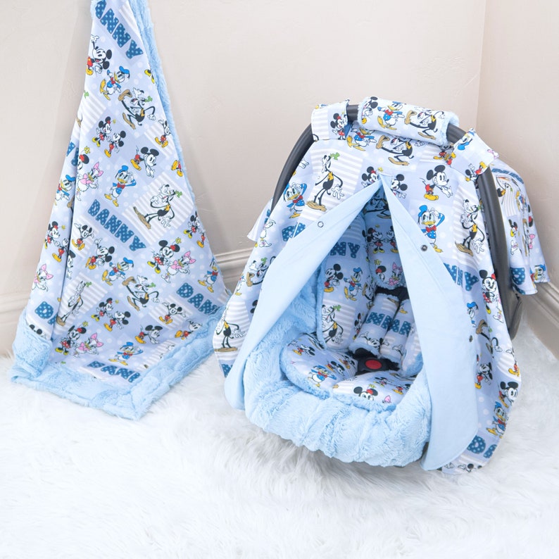 May include: A blue baby car seat cover featuring a Mickey Mouse and friends print. The cover has a hood and a padded insert. The cover is personalised with the name "Danny".