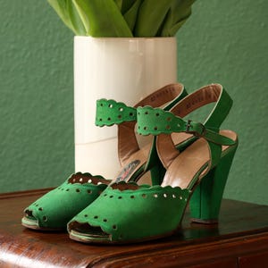 May include: A pair of green suede open-toe heels with scalloped edges and ankle straps. The heels have a chunky design and are set on a wooden surface. A white vase with green leaves is in the background.