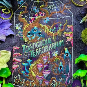 May include: A cross-stitch embroidery on dark fabric featuring a terrarium design with brown tree branches, colorful mushrooms, small blue creatures with eyes, and spiderwebs. The text TOADGHOUL TERRORARIUM is stitched in green and blue letters.