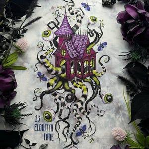 Peut inclure: Broderie au point de croix d'une maison hantée avec un toit et des murs violets, entourée de vignes rayées noires et blanches, d'yeux et de papillons violets. Le texte "13 Eldritch Lane" est brodé en bas. Le motif est encadré de fleurs et de feuillages violets foncés.