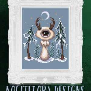 May include: A cross-stitch pattern featuring a whimsical creature with a single eye, antlers, and a crescent moon above its head. The creature is standing in a snowy forest with pine trees and icicles. The pattern is framed in a white ornate frame.