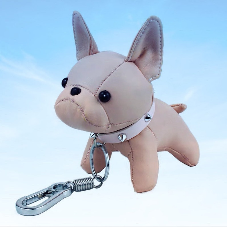 May include: A light pink leather French bulldog keychain with a silver metal ring and a spring clip. The bulldog has a pink studded collar.