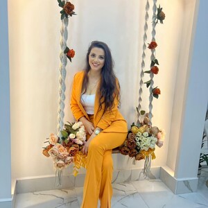 May include: A woman wearing a bright orange pantsuit sits on a macrame swing. She is smiling and looking at the camera. The swing is decorated with artificial flowers.