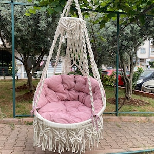 May include: A hanging macrame chair with a round, white frame and a plush, dusty rose cushion. The chair is suspended from a metal frame and features intricate woven detailing and fringe.