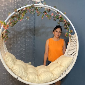 Op de afbeelding: Een witte hangstoel met een crèmekleurig kussen. De stoel is versierd met een krans van groene bladeren en roze bloemen. Een vrouw staat achter de stoel.