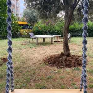 May include: A wooden swing with gray rope hanging from a tree in a backyard. The swing is made of natural wood and has a simple design. The background features a green lawn, a tree, and a table with chairs.