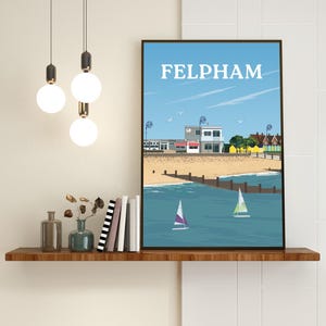 Op de afbeelding: Een ingelijste kunstdruk met een gestileerde illustratie van de kustlijn van Felpham. De afbeelding toont een zandstrand, een houten golfbreker in de blauwe zee, twee zeilboten en kustgebouwen onder een heldere lucht met de tekst FELPHAM.