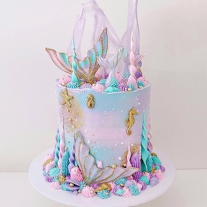 May include: A mermaid-themed cake with pastel blue, purple, and pink frosting. The cake is decorated with gold accents, mermaid tail and seahorse decorations, and edible sea shells. The cake sits on a white cake stand.