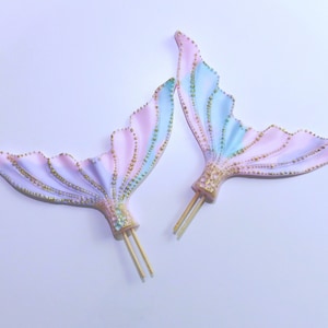 以下が含まれることがあります： 2つの人魚の尾のケーキトッパー。尾はパステルピンク、ブルー、ラベンダーで、金色のアクセントが付いています。各尾には、ケーキやカップケーキに簡単に挿入できる木製のスティックが付いています。