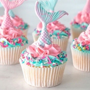 May include: Several cupcakes with mermaid tail toppers. Each cupcake has a light blue frosting base, pink frosting swirls, and colorful sprinkles. The mermaid tails are pink and blue.