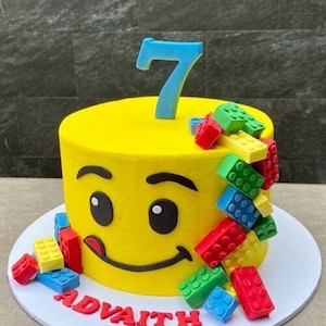 Puede incluir: Una tarta de cumpleaños amarilla decorada con una cara sonriente y caramelos de bloques de construcción de colores. Un adorno azul con el número 7 está centrado en la parte superior. El nombre "ADVAITH" está escrito en letras rojas en la base de la tarta.