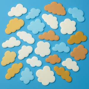 Puede incluir: Una colección de decoraciones en forma de nube en varios colores, incluyendo blanco, azul claro, marrón claro y amarillo mostaza, se dispersan sobre un fondo azul brillante. Las decoraciones probablemente están hechas de un material suave y están dispuestas en un patrón aleatorio.
