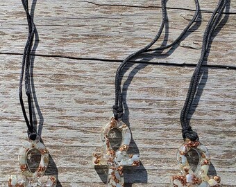 May include: Three Ankh pendant necklaces. Each pendant is a clear resin ankh shape, filled with white and copper-colored fragments. The pendants hang from black cords, displayed on a weathered wooden surface. The ankh is an ancient Egyptian symbol.